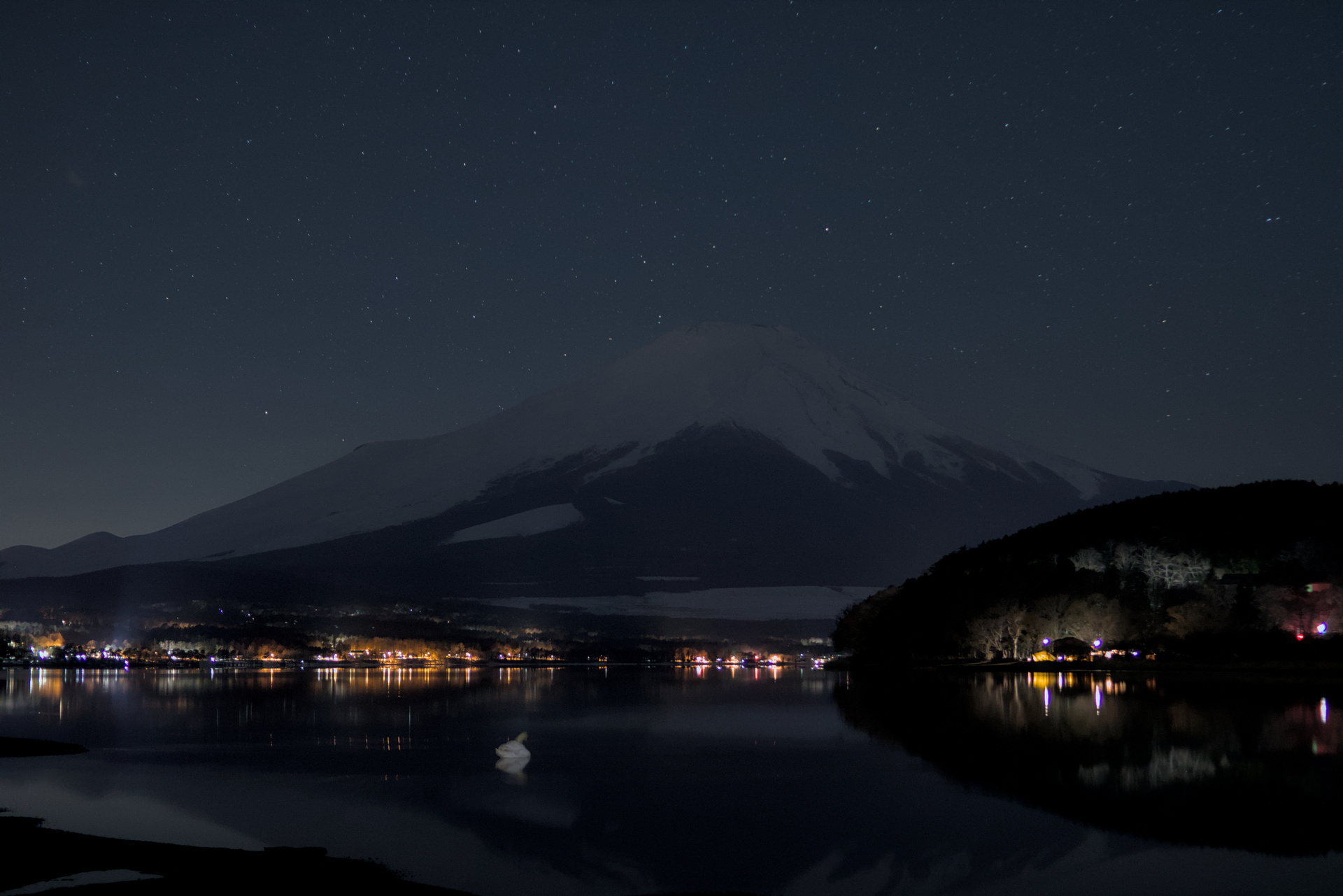 Mount Fuji at night with its reflection in a lake, bright lights at its foot and dimmer stars in the sky.